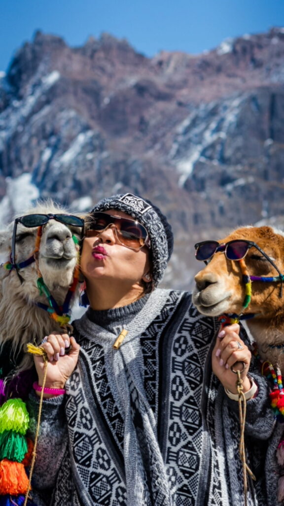 A traveler wearing sunglasses and an Andean poncho posing between two llamas wearing colorful traditional tassels.