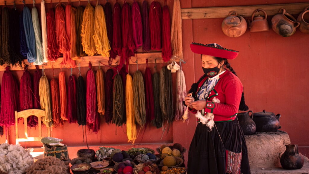 Una mujer con el traje tradicional de Chinchero de pie frente a estantes llenos de lana colorida teñida de forma natural.