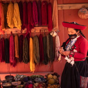 Una mujer con el traje tradicional de Chinchero de pie frente a estantes llenos de lana colorida teñida de forma natural.