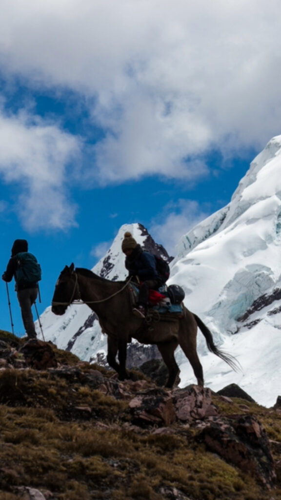 A person riding a horse up a steep, rocky mountain trail with a massive snow-capped glacier in the background.