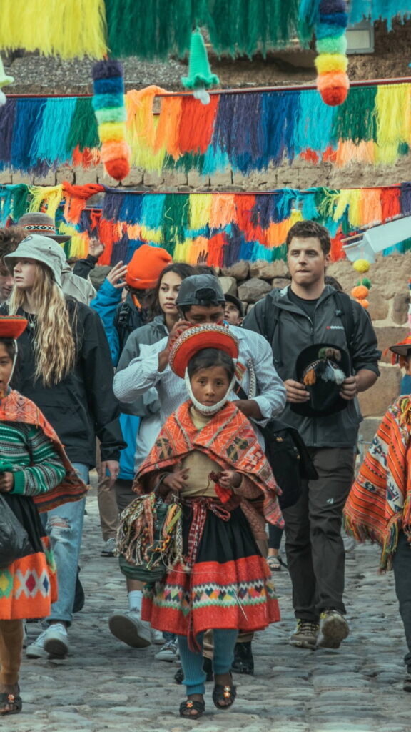 Una niña joven con vestimenta tradicional andina colorida caminando entre turistas bajo coloridos estandartes colgantes.