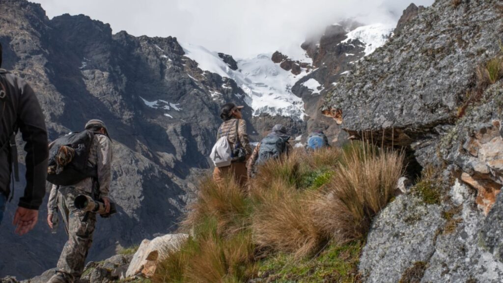 Un grupo de exploradores en una caminata de montaña ascendiendo por un sendero rocoso andino hacia un glaciar masivo cubierto de nieve bajo un cielo nublado.