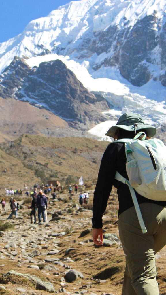 Un excursionista con una mochila caminando por un sendero de montaña rocoso hacia un gran pico cubierto de nieve.