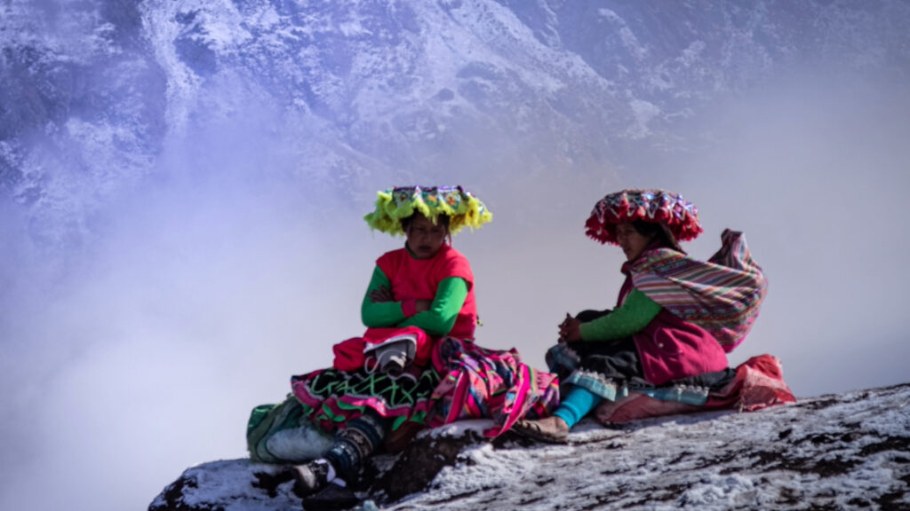 Two local Andean women in traditional vibrant red and green clothing sitting on a snowy mountain ridge surrounded by mist.