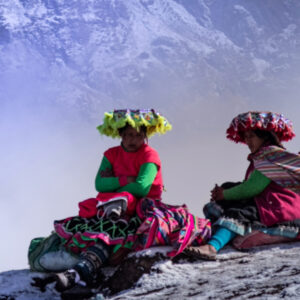 Two local Andean women in traditional vibrant red and green clothing sitting on a snowy mountain ridge surrounded by mist.