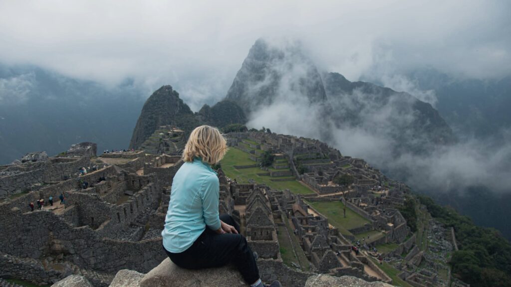 Un viajero se sienta en un saliente de piedra con vista a las antiguas ruinas de Machu Picchu, con el pico de la montaña Huayna Picchu parcialmente cubierto por las nubes matutinas.