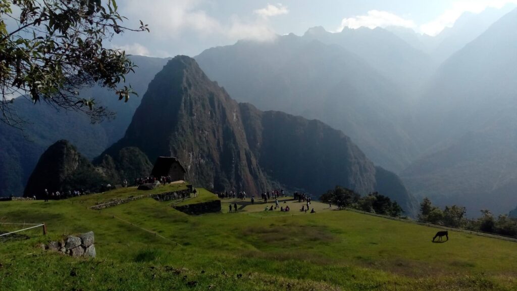 Un guía de turismo profesional mostrando las ruinas de piedra de Machu Picchu a un grupo de viajeros bajo un cielo azul brillante con la montaña Huayna Picchu detrás.