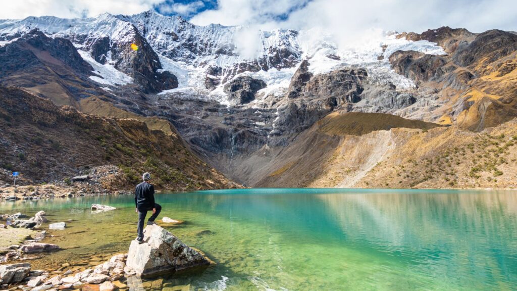 Un viajero de pie sobre una roca con vistas a la laguna turquesa Humantay, con un enorme glaciar cubierto de nieve al fondo.