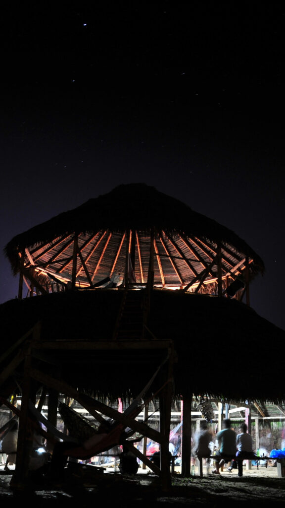 Una maloca tradicional amazónica de madera iluminada desde el interior contra un cielo nocturno oscuro en Pillcopata.