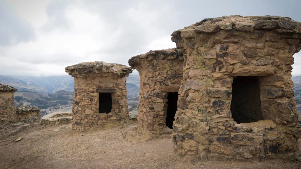 Una hilera de antiguas torres funerarias de piedra de Ninamarca, conocidas como chullpas, situadas en la ladera de una montaña que domina un valle bajo un cielo andino nublado.