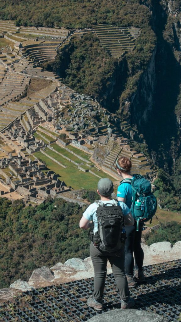 Dos viajeros con mochilas parados sobre una terraza de piedra antigua, mirando hacia abajo la ciudadela de Machu Picchu y la montaña Huayna Picchu.