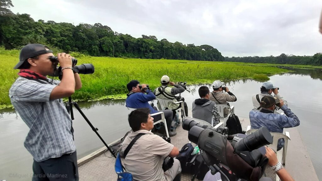 Un grupo de observadores de aves de Pichitankas en una balsa de fondo plano, utilizando binoculares y cámaras profesionales para avistar la vida silvestre a lo largo de una exuberante orilla de río tropical bajo un cielo nublado.