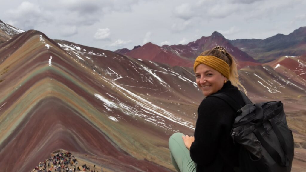 Una excursionista sonriente con una mochila se sienta frente a las vibrantes franjas minerales de múltiples colores de la Montaña de Siete Colores, bajo un cielo nublado en los Andes peruanos.