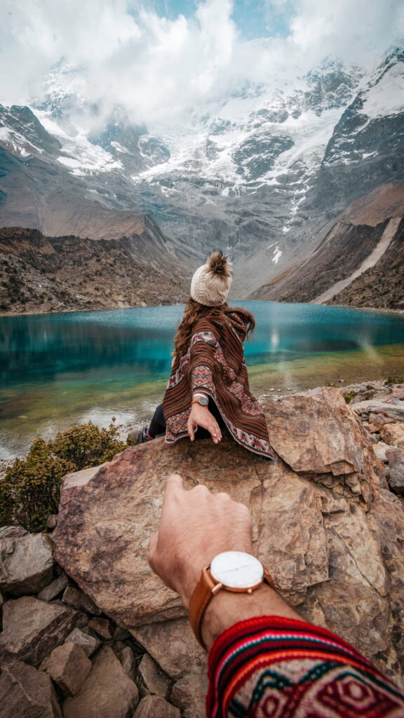 Un primer plano de una mano acercándose a una apacheta de piedra (montículo de ofrenda) con vistas a las aguas turquesas de la laguna Humantay.