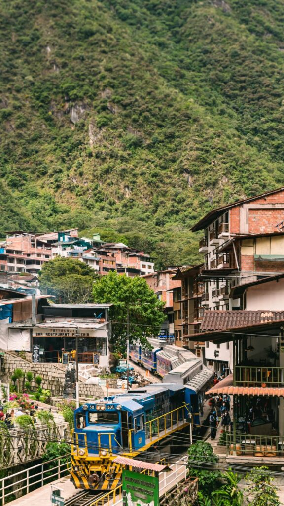 Un tren turístico azul estacionado en las vías, rodeado de las escarpadas y exuberantes montañas verdes y los coloridos edificios de Aguas Calientes.