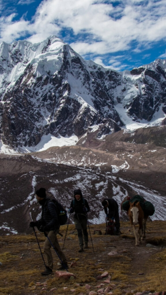 A group of hikers with trekking poles walking through a wide, rocky valley toward a towering snow-covered mountain peak.