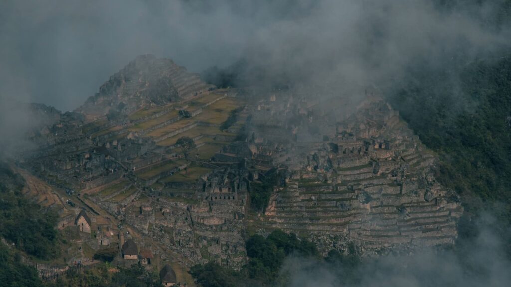 Una vista aérea atmosférica de las ruinas de piedra y las terrazas agrícolas de Machu Picchu parcialmente ocultas por una densa y arremolinada neblina de montaña y nubes.
