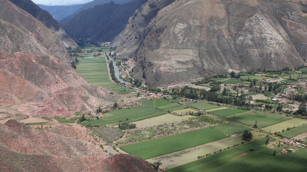 Vista aérea panorámica de los exuberantes campos verdes y el río serpenteante del Valle Sagrado, rodeado por las majestuosas y secas laderas de los Andes peruanos.