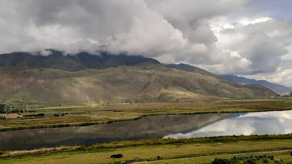 Una vista panorámica de la Laguna de Huacarpay reflejando densas nubes blancas y las montañas andinas circundantes de gran altitud en su superficie de agua oscura y tranquila.