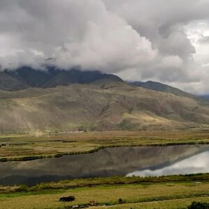 Una vista panorámica de la Laguna de Huacarpay reflejando densas nubes blancas y las montañas andinas circundantes de gran altitud en su superficie de agua oscura y tranquila.